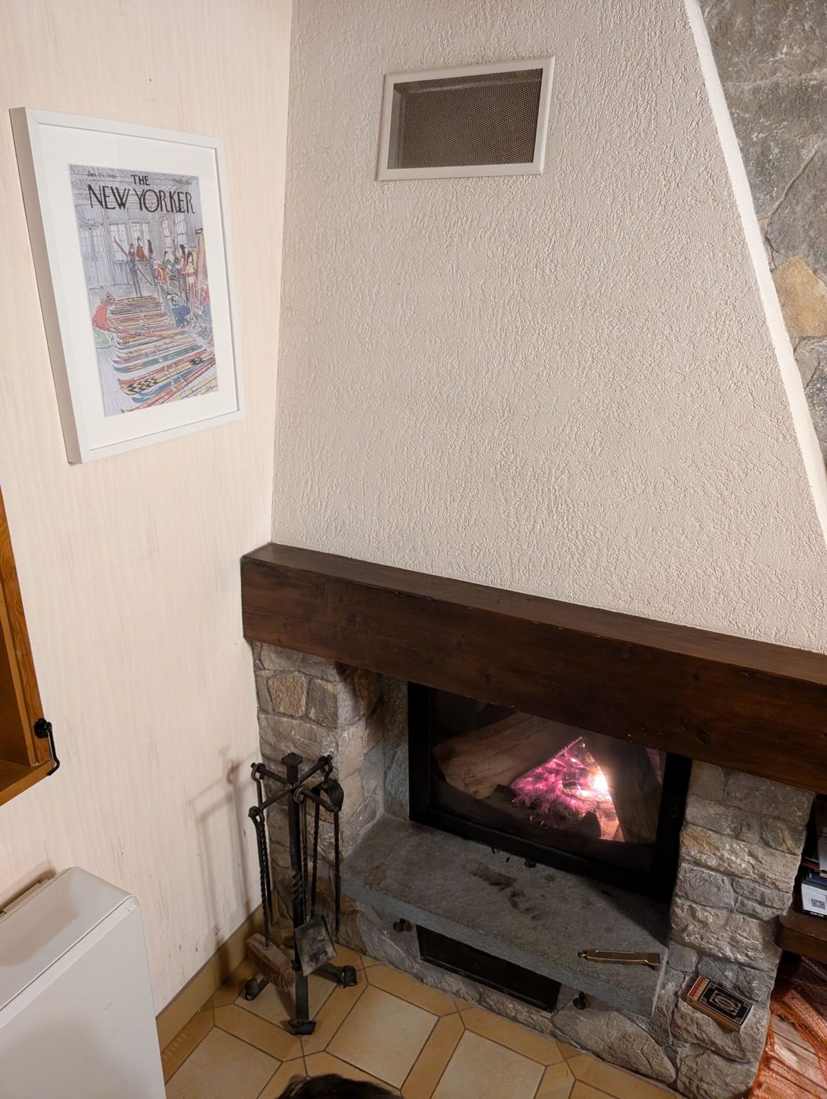 The stone fireplace with the wood burner lit, and a framed New Yorker cover on the wall beside it
